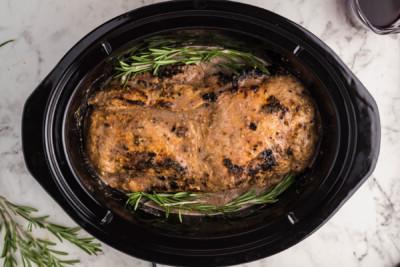 Lombo di maiale posizionato nel slow cooker, immerso nel ricco sugo aromatico e guarnito con rametti di rosmarino fresco, prima dell'inizio della cottura.