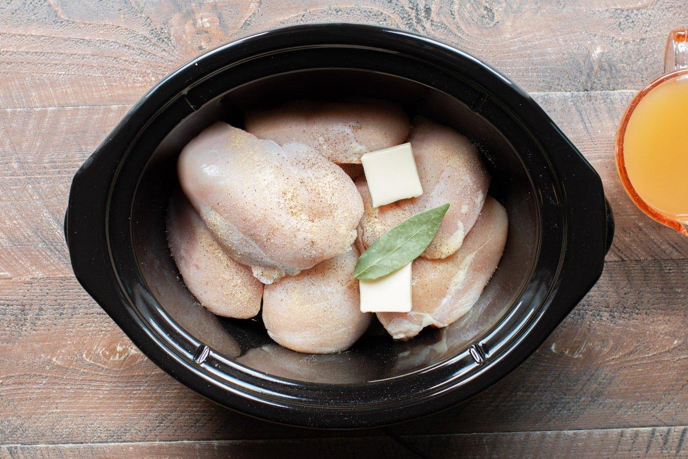 Pollo sfilacciato in slow cooker 5 petti di pollo crudi in una pentola a cottura lenta ovale con fiocchi di burro, foglia di alloro e pepe sopra.