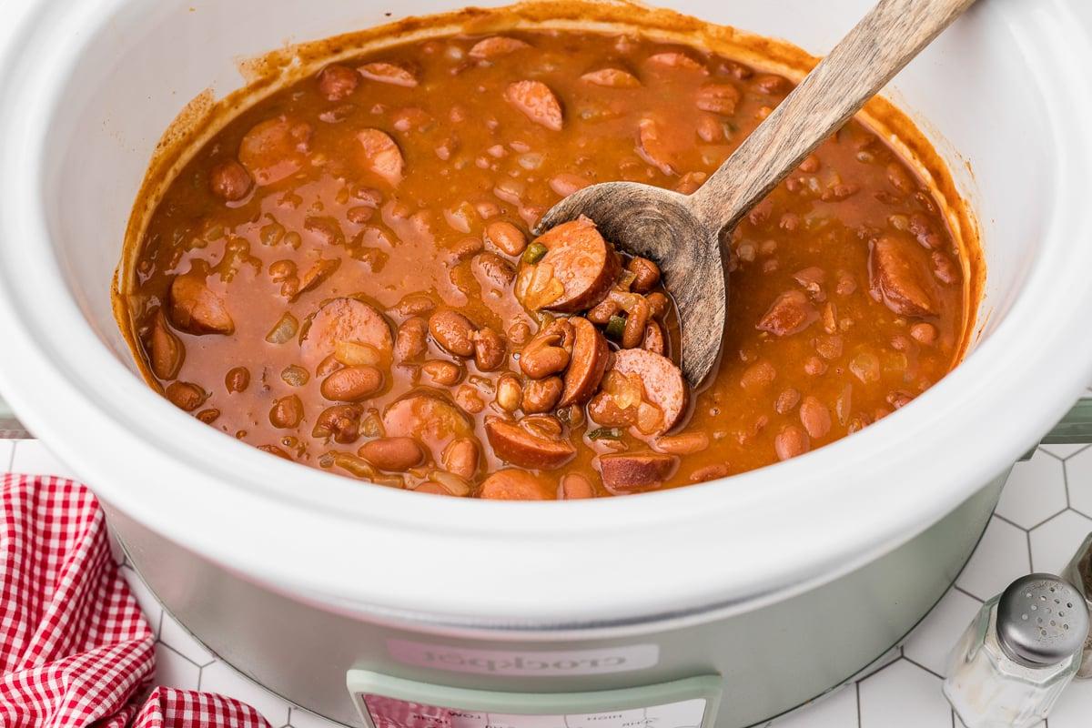 Fagioli e salsicce in una slow cooker con un cucchiaio di legno all'interno.