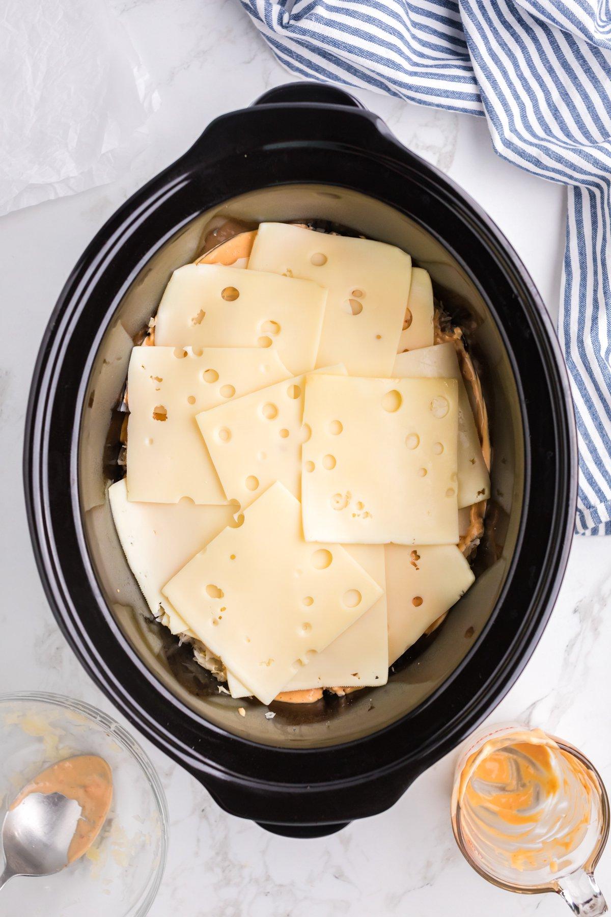 Casseruola di Pollo alla Reuben a Cottura Lenta 7 Formaggio svizzero aggiunto sopra pollo, crauti e salsa nella slow cooker.