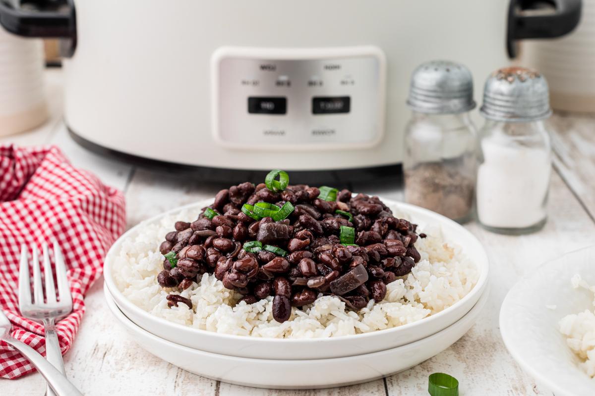 Fagioli Neri Cottura Lenta 7 Fagioli neri serviti su un piatto con riso, in primo piano, con una slow cooker sullo sfondo.