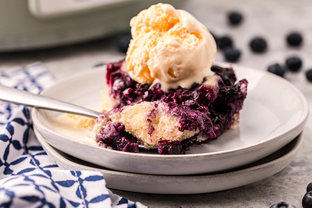 **Cobbler ai Mirtilli della Slow Cooker** 7 Una forchetta che tiene un morso di cobbler di mirtilli al slow cooker su un piatto con il resto della porzione.