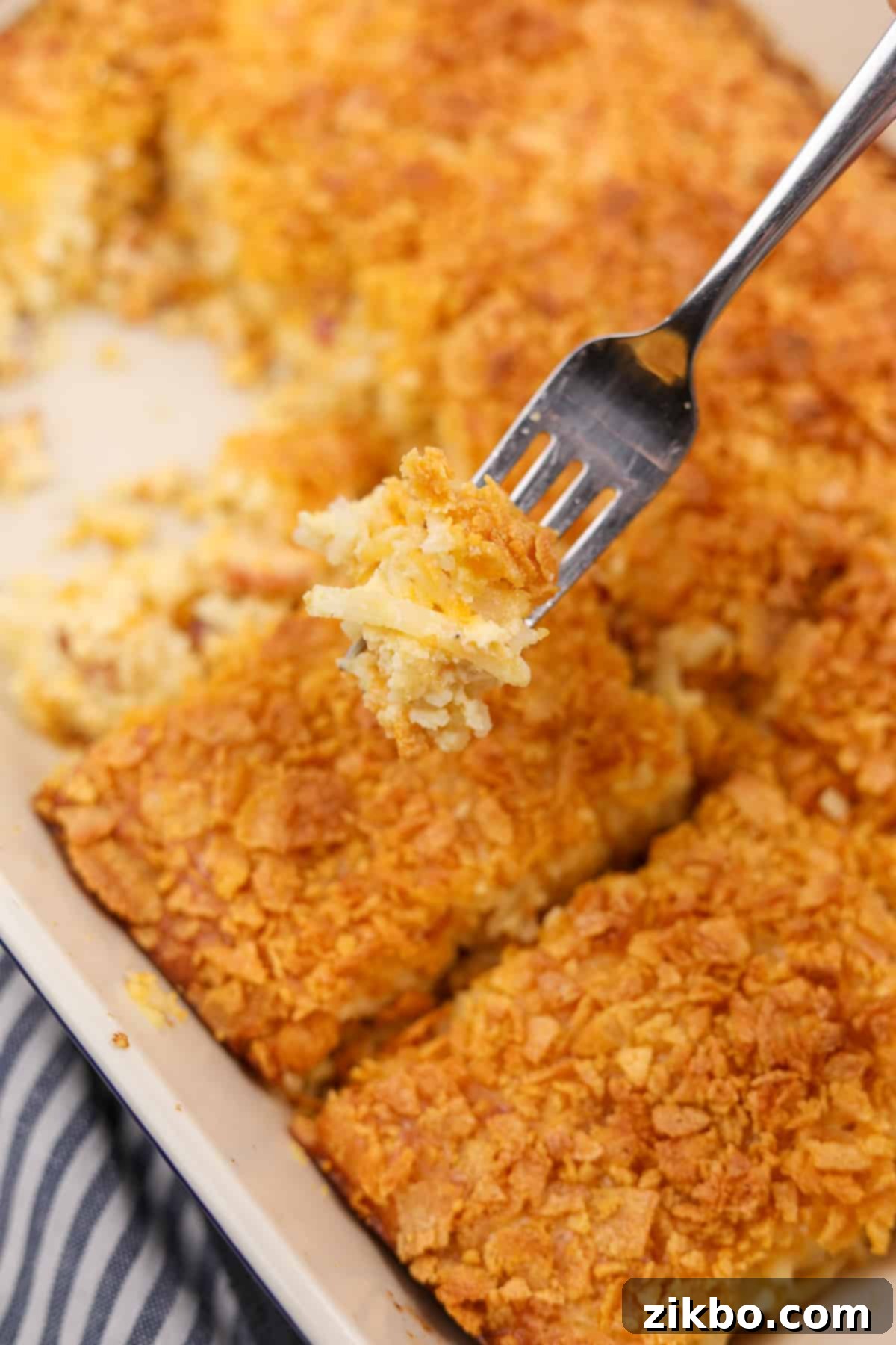 Ultimate Breakfast Hash Brown Bake 8 A fork showing the texture of a shredded hashbrown casserole, which is in the background and topped with crunchy cornflakes.