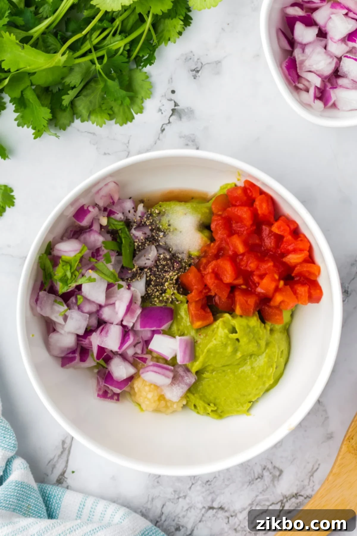 Robust Chunky Guacamole 4 A bowl of mashed avocado topped with red onion, tomato, garlic, pepper, salt, and cilantro, ready for mixing.
