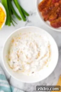 A white bowl filled with a sour cream based dip, with bowls of cooked bacon and cheese on the side, chilling in the refrigerator.