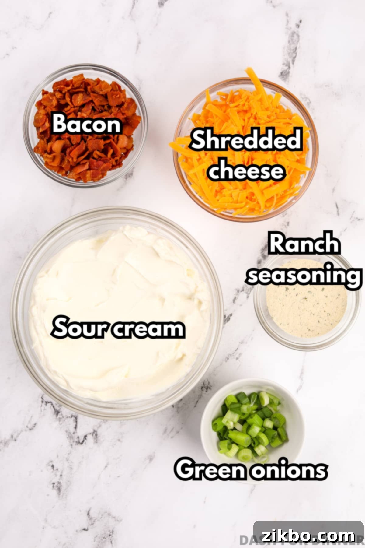 Ultimate Loaded Baked Potato Dip 3 An overhead view of bowls of fresh ingredients, including sour cream, shredded cheddar cheese, crumbled bacon, ranch seasoning, and chopped green onions, ready for mixing.