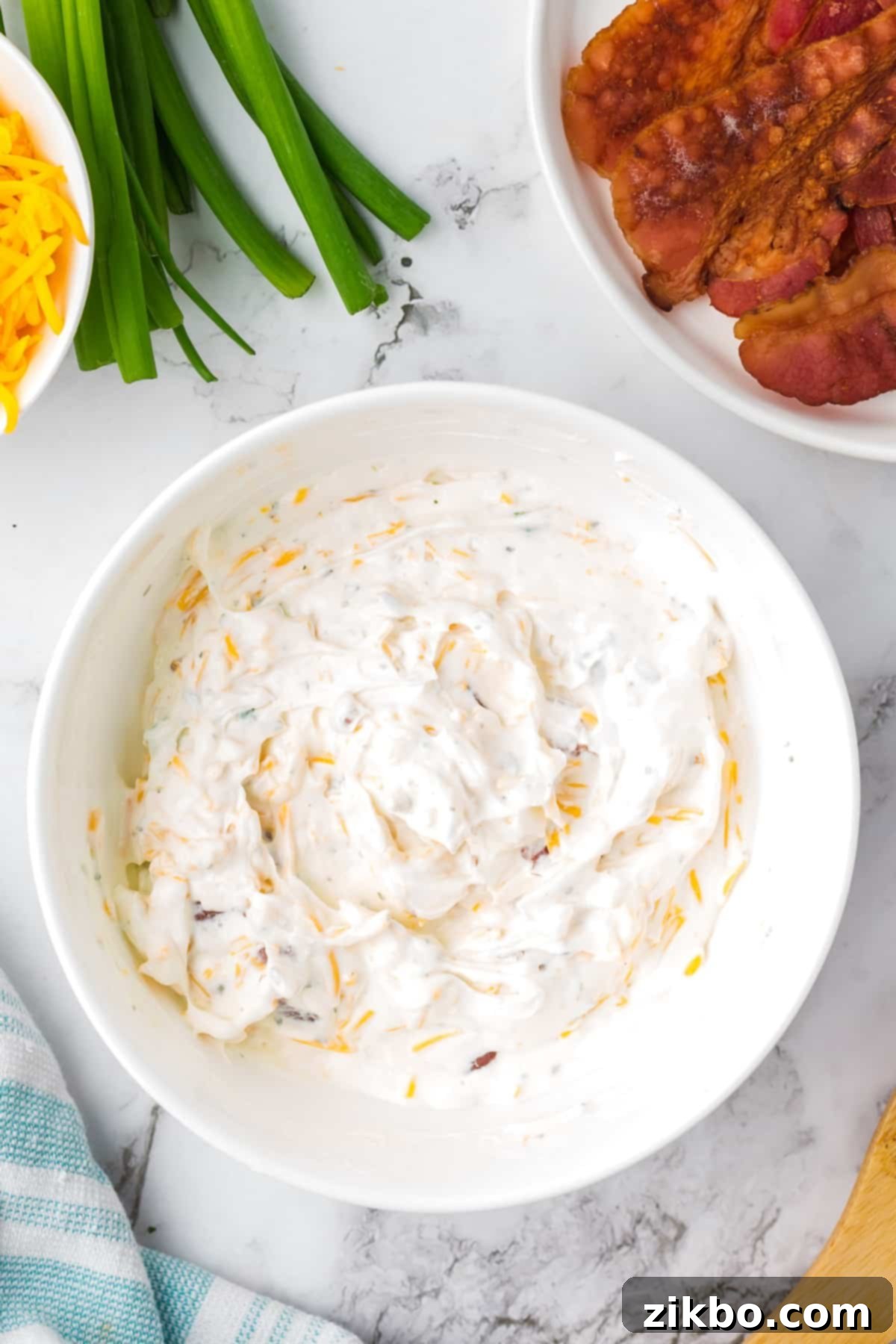 Ultimate Loaded Baked Potato Dip 5 A white bowl filled with a sour cream based dip, with bowls of cooked bacon and cheese on the side, ready for chilling.
