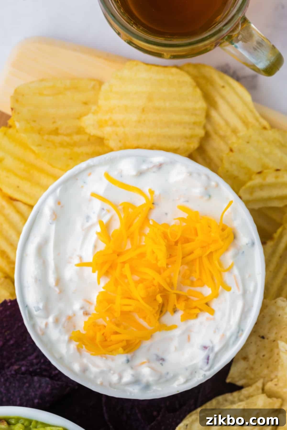 A bowl of creamy beer cheese dip topped with shredded cheddar cheese, with ruffled potato chips on the side for dipping.