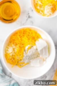 A bowl filled with softened cream cheese, shredded cheeses, and ranch seasoning, with a small measuring cup of beer in the top left corner.
