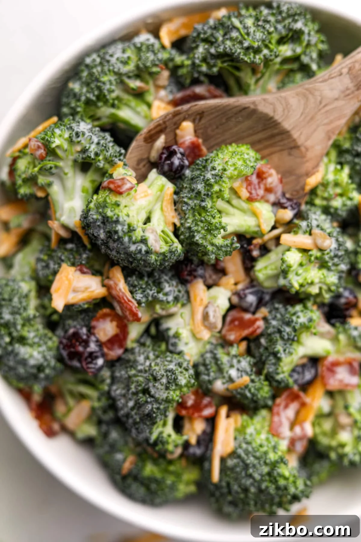 Crunchy Broccoli Delight 2 A wooden spoon taking a small portion from a large bowl of broccoli salad. It's garnished with bacon and cheddar cheese shreds, showcasing its rich texture and appealing colors.
