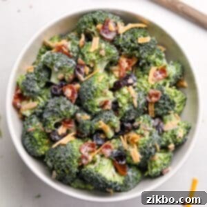 A white bowl filled with broccoli salad. It's garnished with cheddar cheese shreds and bacon.
