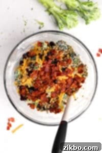 A bowl of broccoli salad with bacon and cheese mixed in, along with cranberries and sunflower seeds, ready for a final stir.