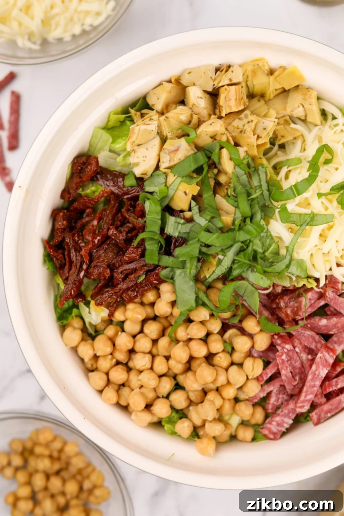 A large bowl of the famous La Scala Chopped Salad, generously topped with sections of golden chickpeas, vibrant sundried tomatoes, fresh basil leaves, tender artichoke hearts, creamy mozzarella, and savory salami slices.