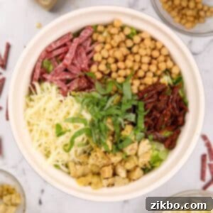 A bowl of vibrant chopped salad, featuring creamy mozzarella cheese, tender artichokes, sweet sun-dried tomatoes, golden chickpeas, savory salami, and fresh basil leaves, all perfectly mixed.