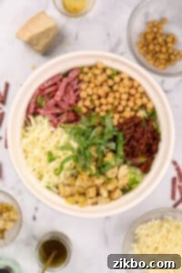 A large salad bowl filled with ingredients, including sections of toppings: mozzarella, artichokes, basil, sun dried tomatoes, chickpeas, and salami on lettuce.