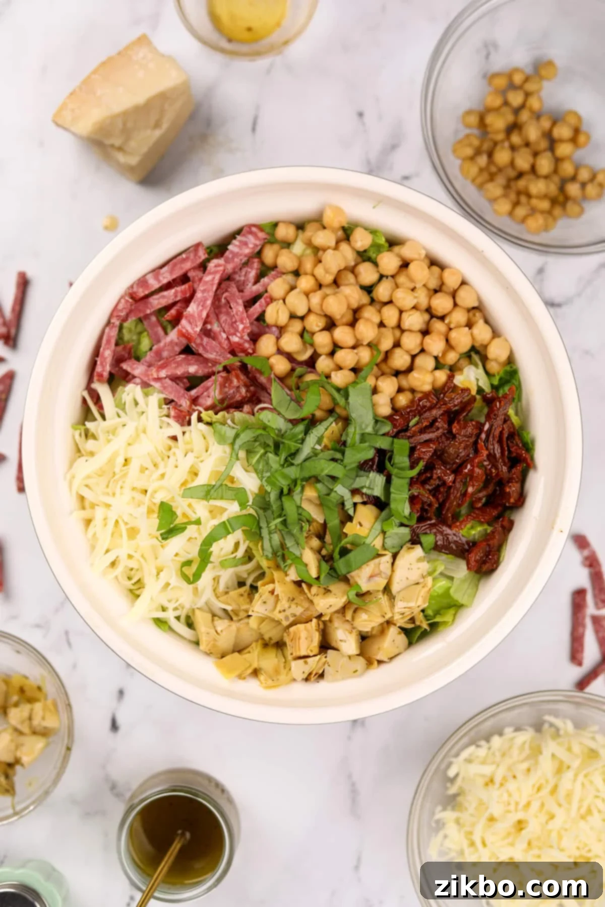 A large salad bowl artfully arranged with various toppings in distinct sections: creamy mozzarella, chopped artichokes, fresh basil, vibrant sun-dried tomatoes, golden chickpeas, and savory salami slices, all resting on a bed of crisp lettuce.