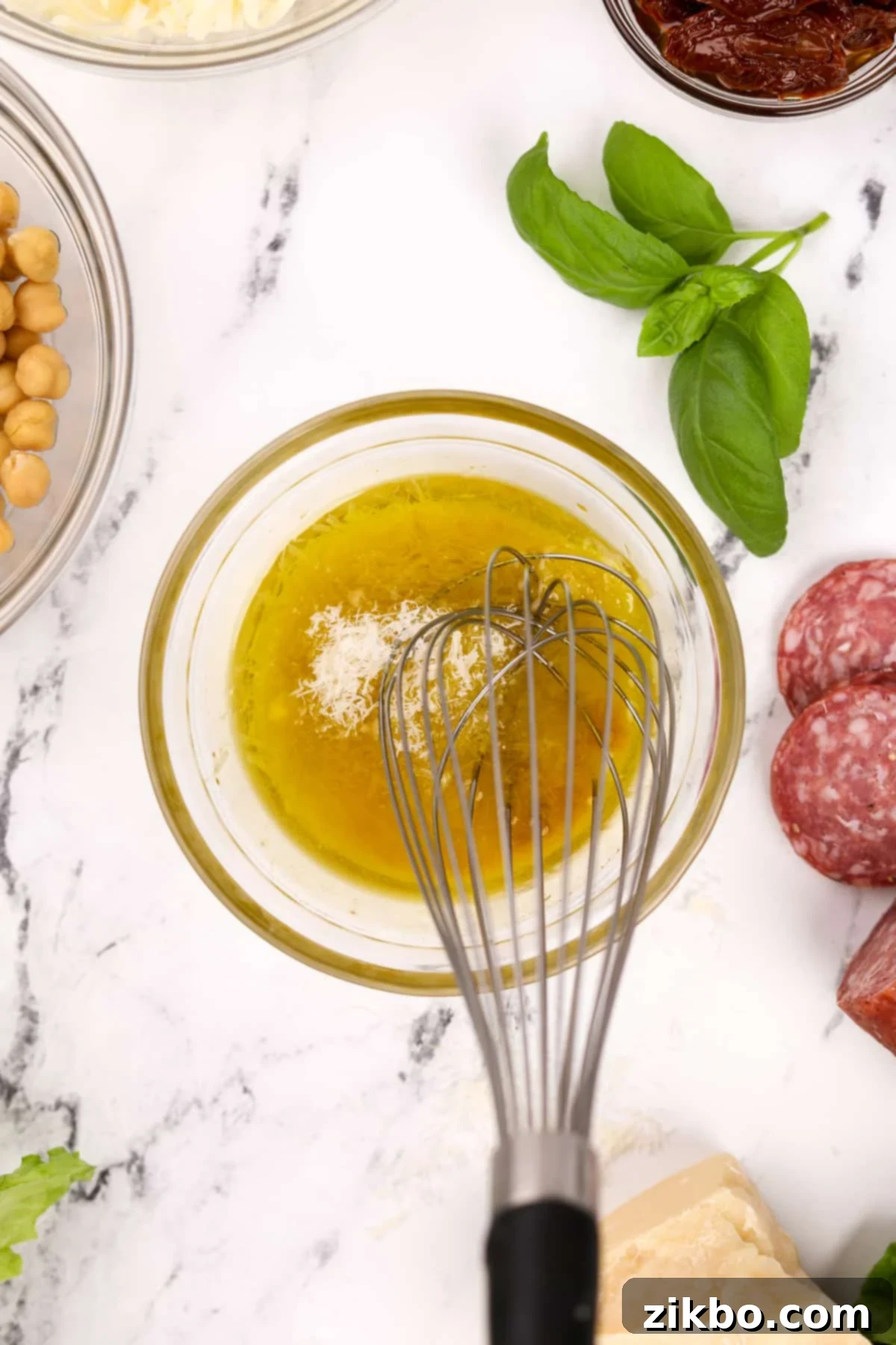 A glass bowl with a whisk, mixing together ingredients for the Leon Dressing, prominently featuring golden olive oil, grated parmesan cheese, and other dressing components.