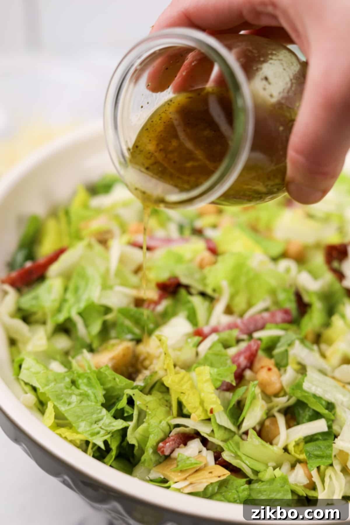 A steady stream of amber-colored salad dressing being poured over a vibrant, large bowl of chopped salad, ensuring even coating.