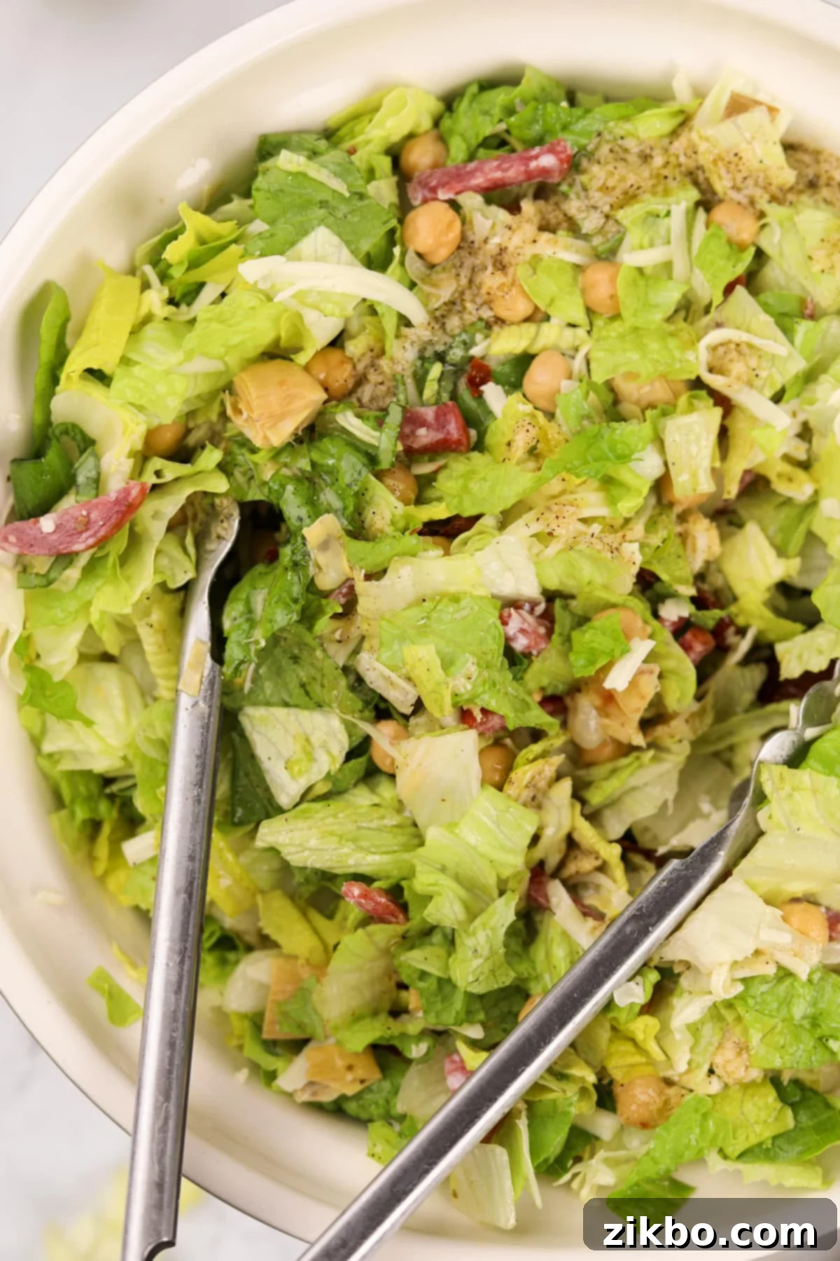 A pair of stainless steel tongs actively tossing a colorful chopped salad in a large bowl, ensuring the dressing is evenly distributed among all ingredients.