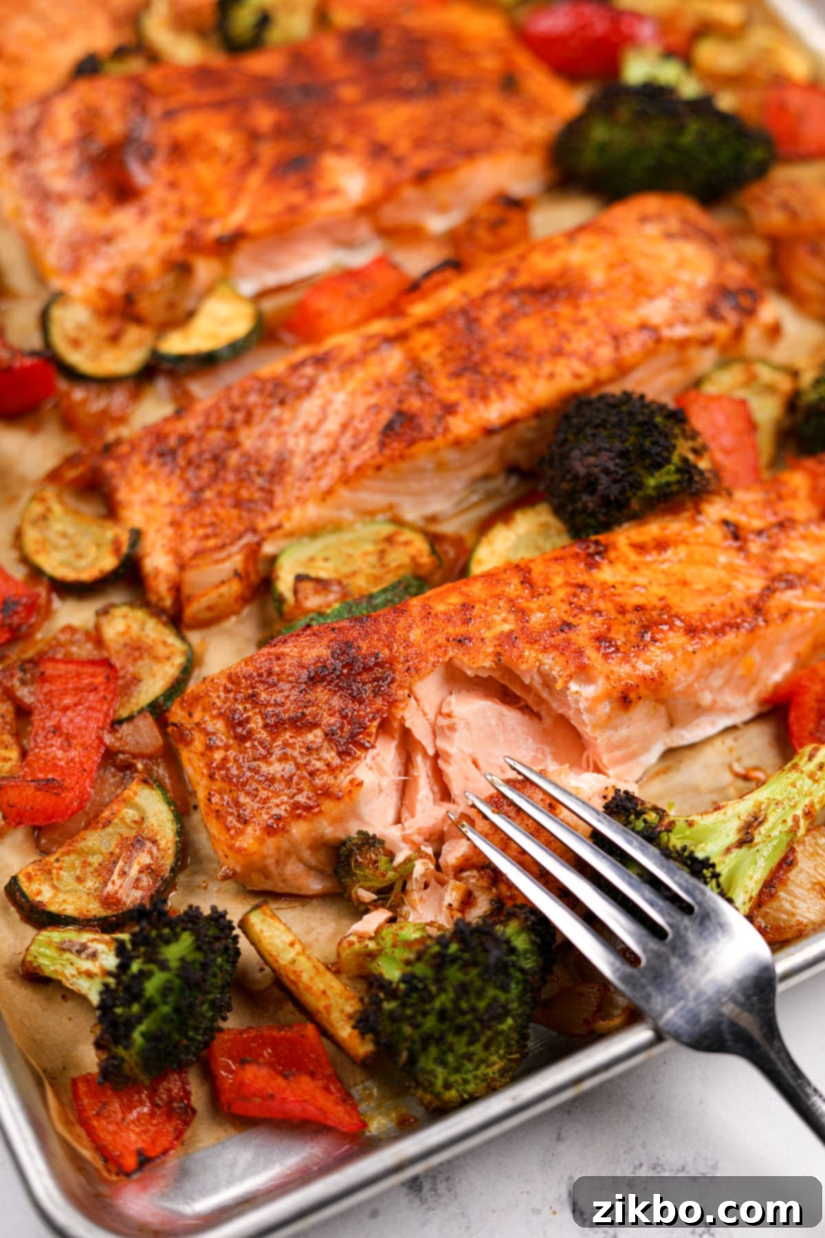 Effortless Sheet Pan Salmon Feast 2 A fork flaking a piece of perfectly cooked salmon on a baking sheet, surrounded by beautifully roasted vegetables including bright green broccoli, red bell pepper, and zucchini.