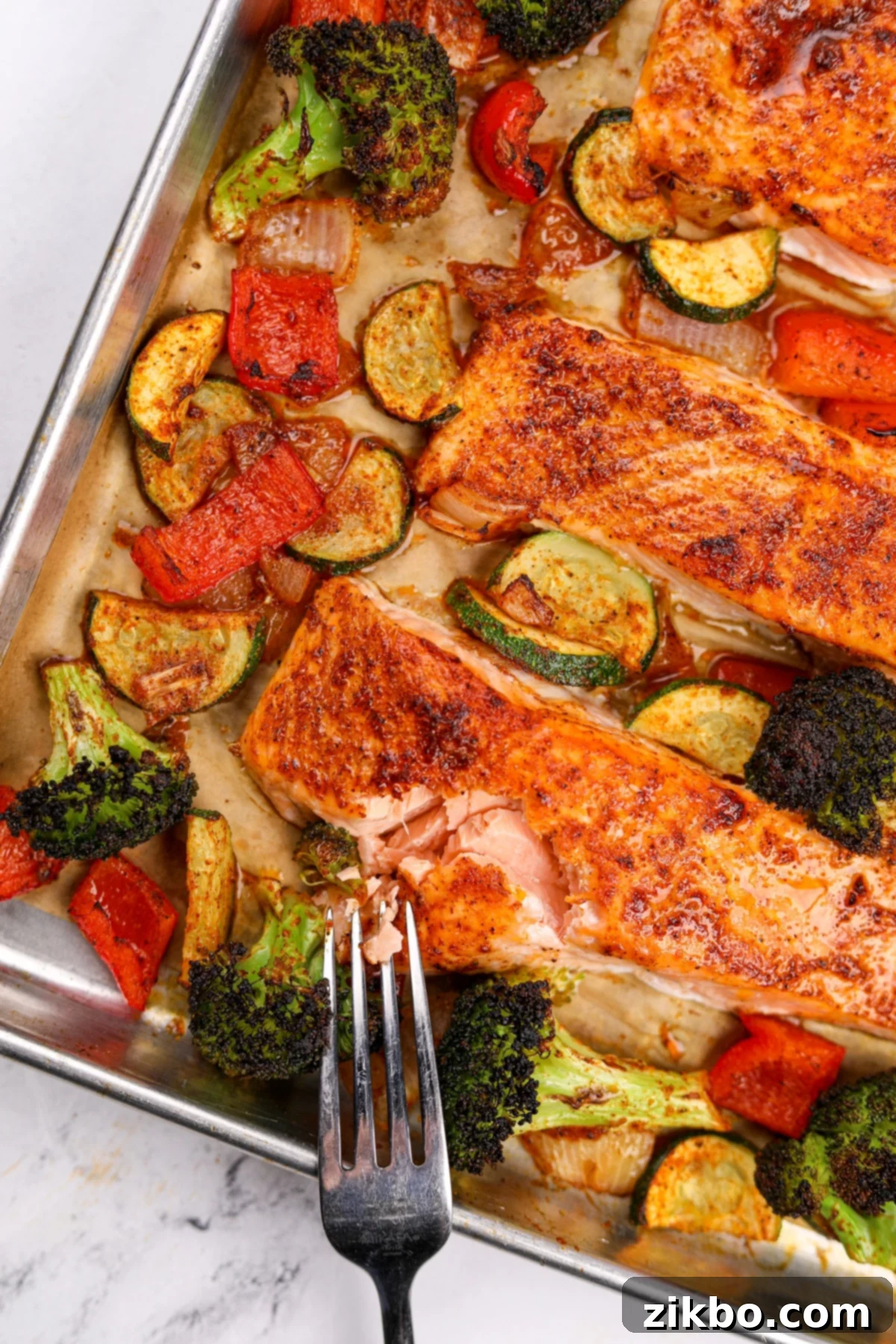Effortless Sheet Pan Salmon Feast 11 A close-up shot of a fork expertly flaking a succulent fillet of roasted salmon, with a colorful medley of perfectly cooked broccoli, red bell pepper, and zucchini visible in the background on the sheet pan.
