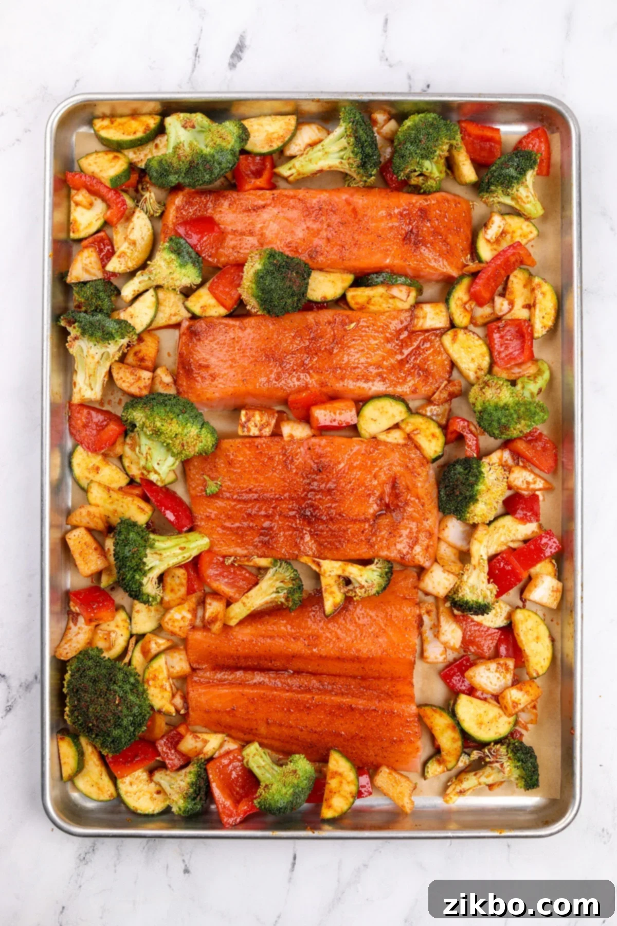 Effortless Sheet Pan Salmon Feast 8 A sheet pan arranged with four seasoned salmon fillets and an assortment of raw, seasoned broccoli, bell pepper, and zucchini spread around them, ready for the oven.