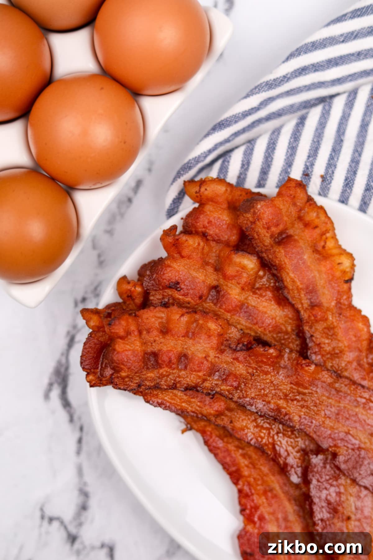 Perfectly Crispy Baked Bacon 7 Crispy bacon slices on a white plate, with eggs on the edge of the frame.
