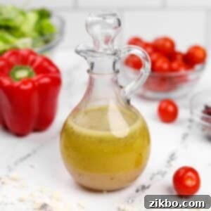 A glass cruet filled with homemade Greek salad dressing, with vibrant red pepper, juicy tomatoes, and crisp lettuce forming a fresh salad in the background.
