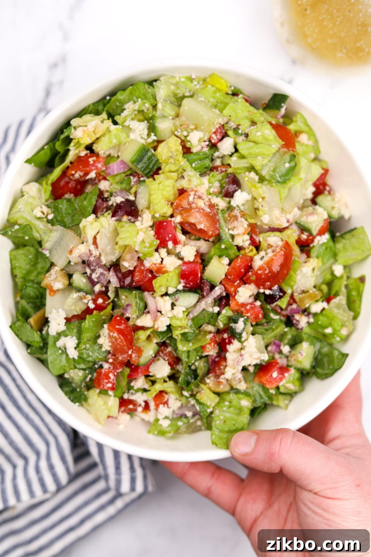 The Perfect Greek Chopped Salad 7 A hand holding a clear glass bowl filled with a fresh, colorful Greek chopped salad, showcasing the vibrant ingredients.