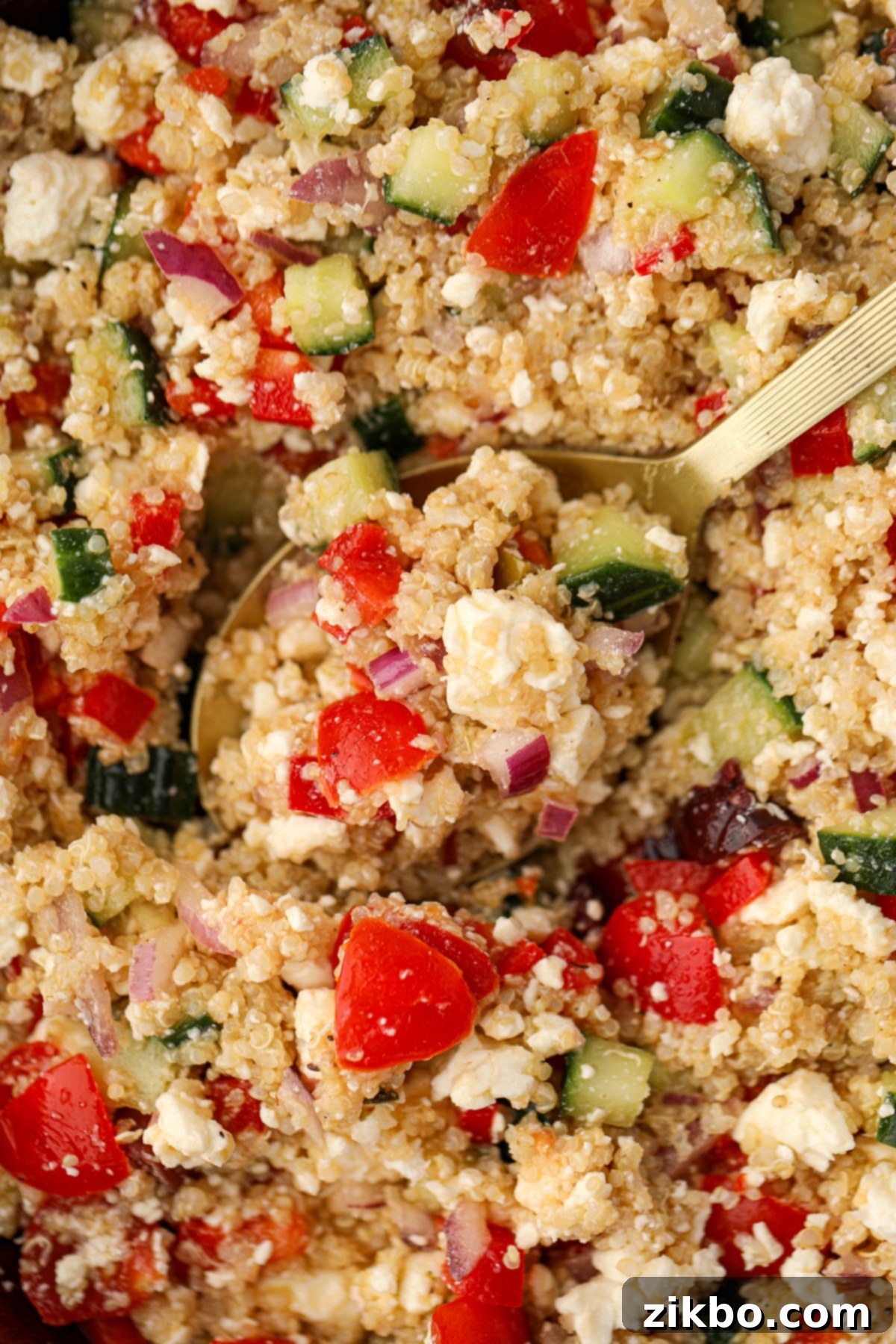 A spoon scooping into quinoa salad featuring red pepper, feta, and cucumbers.