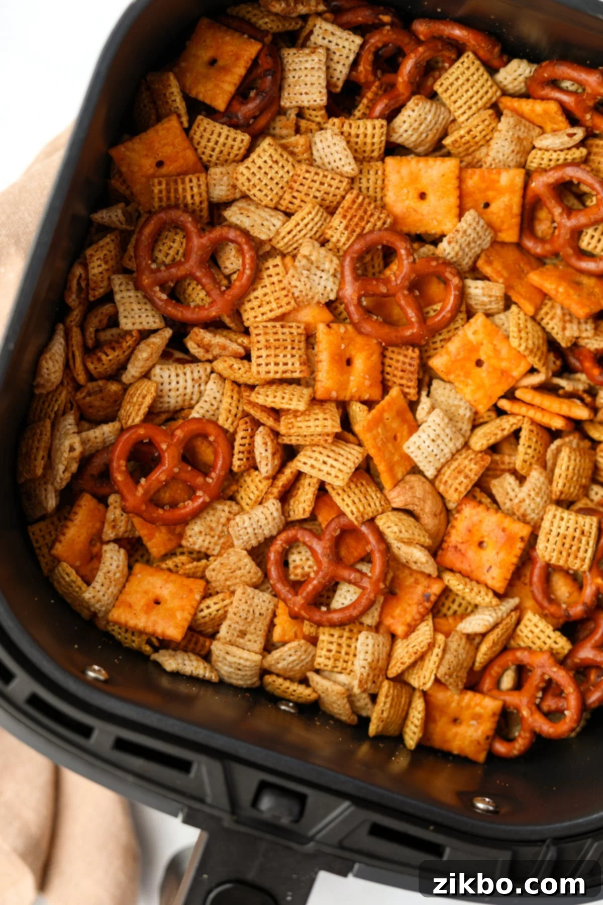 An air fryer basket filled with snack mix featuring pretzels, cereal, and cheez-its.