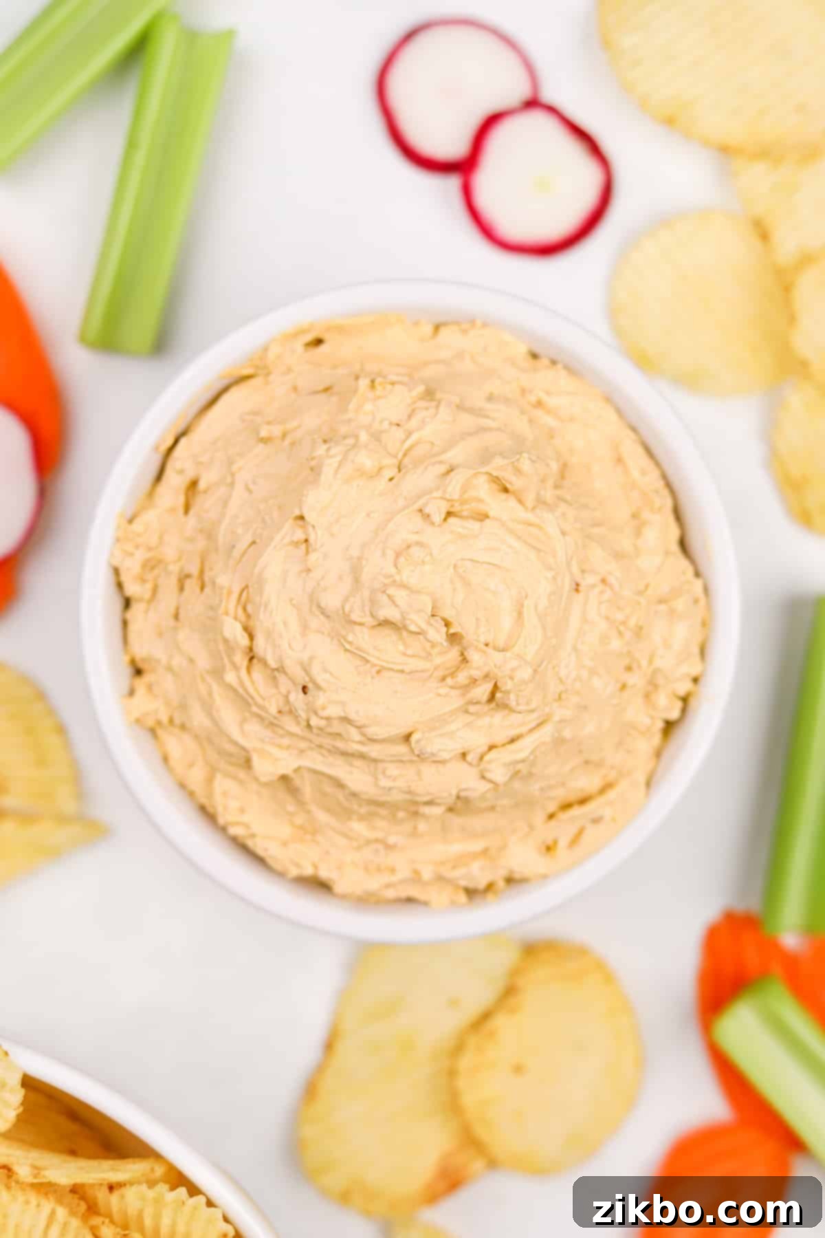 A vibrant bowl of homemade French onion dip is positioned centrally on a platter, artfully surrounded by a selection of crunchy potato chips and an assortment of fresh, colorful vegetables including carrots, celery, and bell pepper slices, creating an inviting spread.