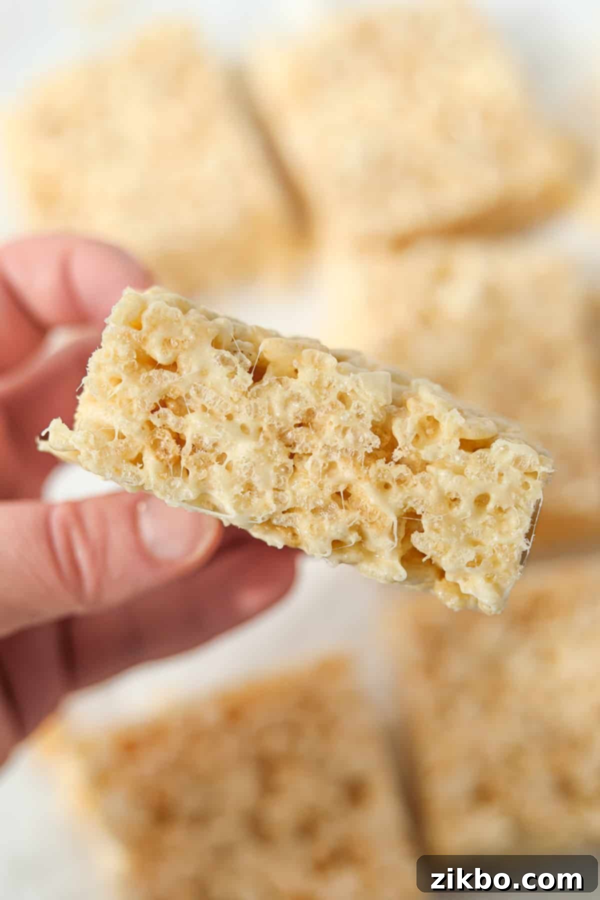 Crispy Sweet Squares 13 A hand holding up a single, perfectly textured Rice Krispie treat, showing off its irresistible chewiness and light, airy structure against a white background.