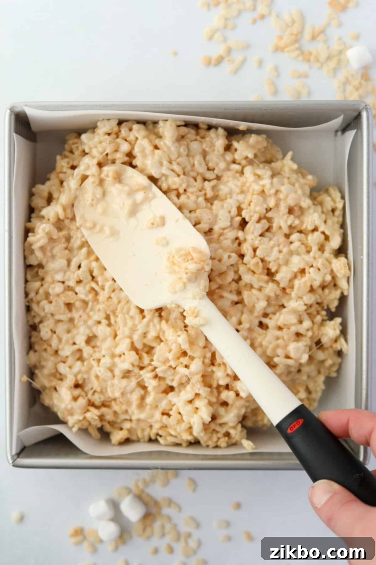 Crispy Sweet Squares 8 The combined cereal and marshmallow mixture being evenly spread into a parchment-lined square baking pan with a spatula.