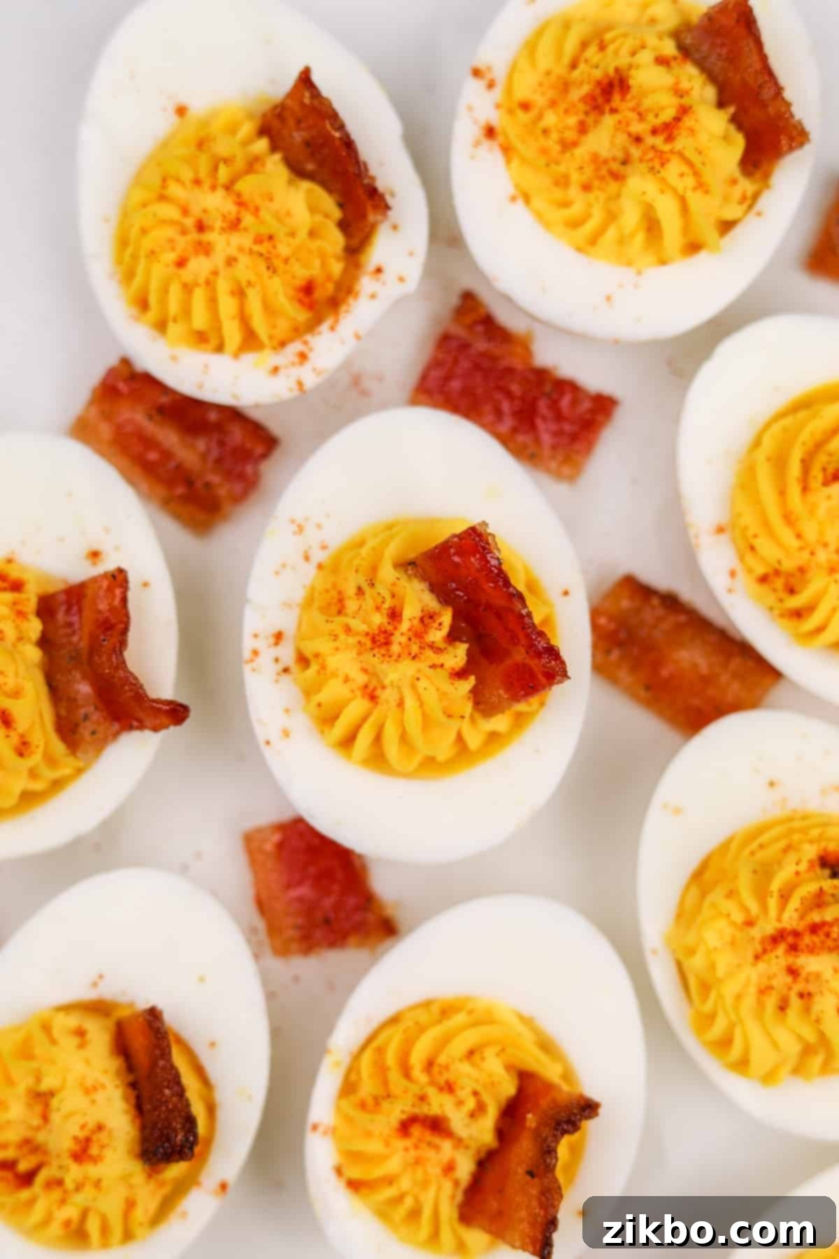 Eight perfectly garnished deviled eggs, each topped with a square of crispy candied bacon and a sprinkle of paprika, arranged beautifully on a serving platter.