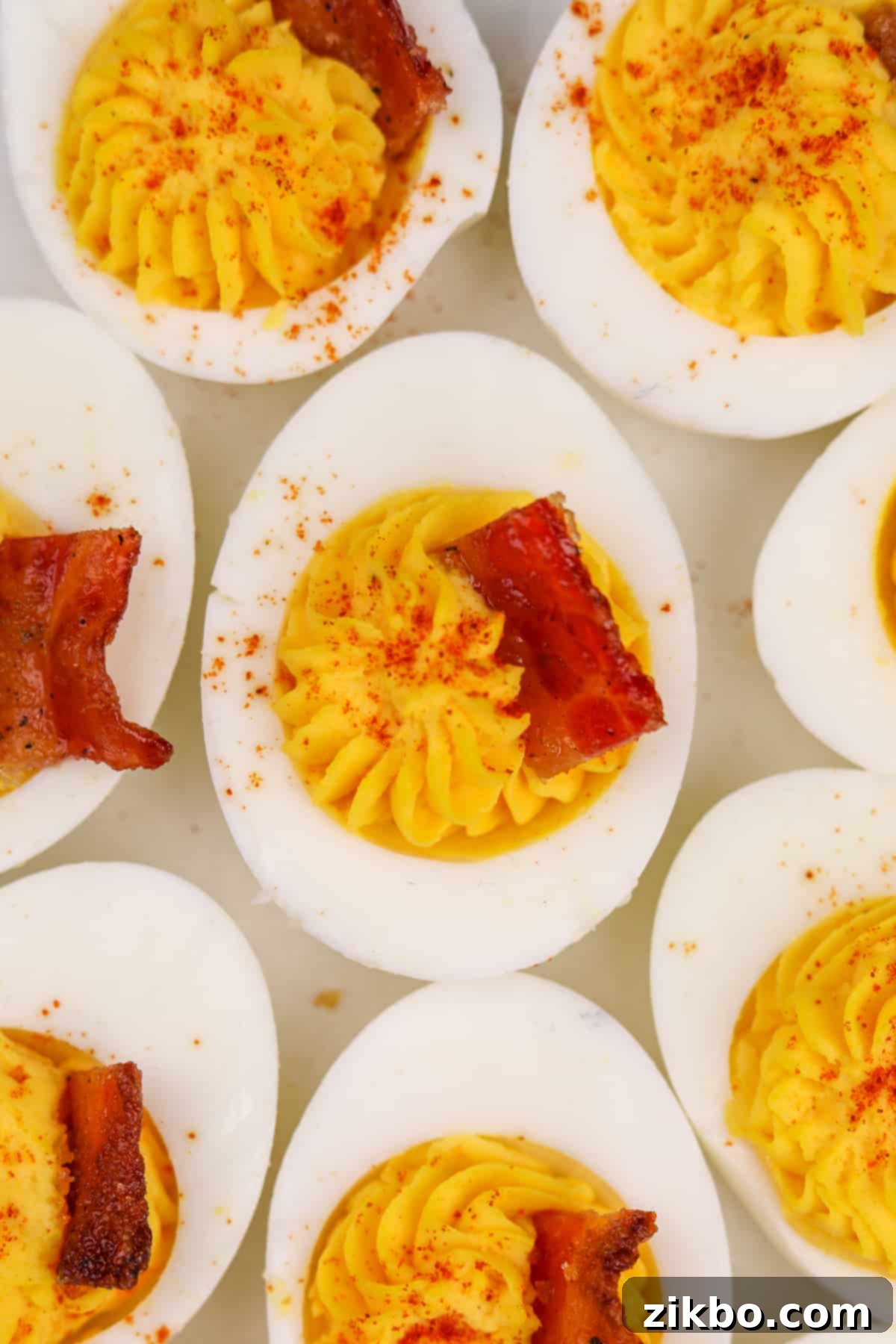 Eight deviled egg halves beautifully arranged on a serving plate, each topped with a cube of candied bacon and a sprinkle of paprika.