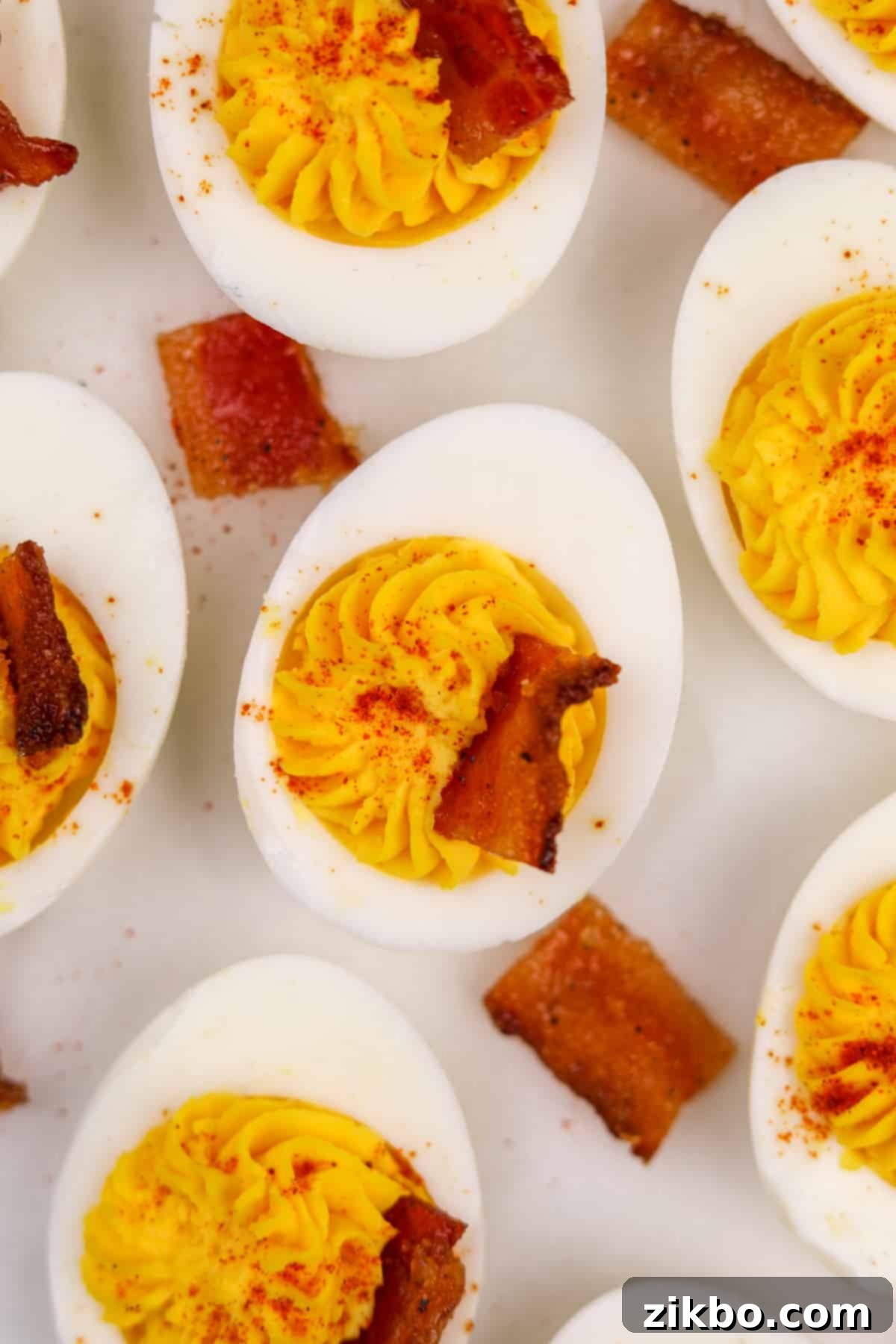 A single deviled egg half, artfully presented with a wedge of glistening candied bacon and a light dusting of paprika, on a neutral background.