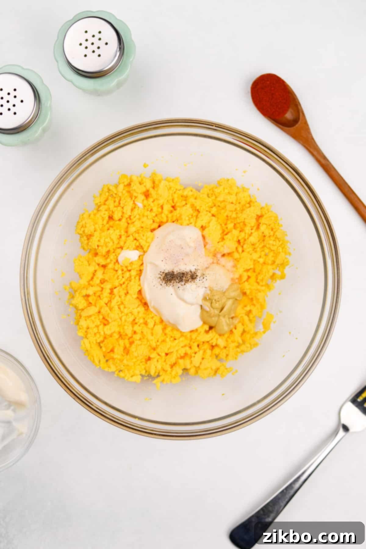 Mayonnaise and a blend of seasonings being added to a bowl of mashed egg yolks, ready for mixing.
