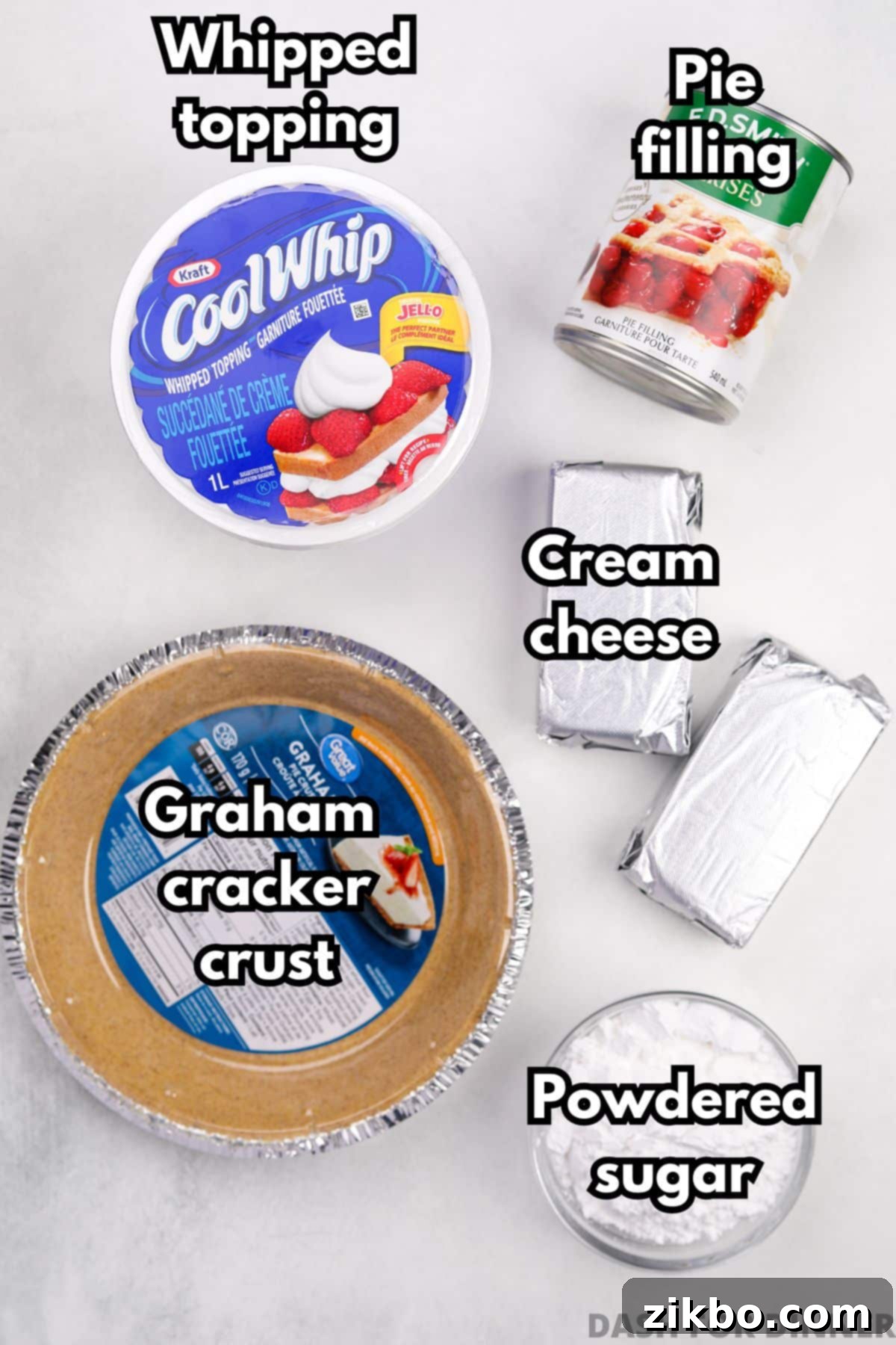 The five essential ingredients laid out for making no-bake cheesecake: a pre-made graham cracker crust, Cool Whip, cherry pie filling, cream cheese, and powdered sugar.