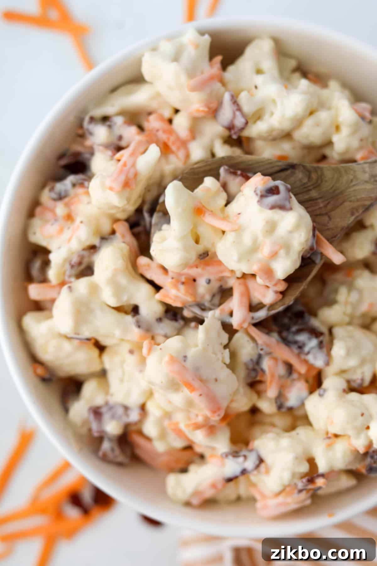 A wooden spoon scooping some cauliflower salad from a bowl, showcasing the creamy dressing, cauliflower florets, bacon bits, and shredded cheddar cheese.