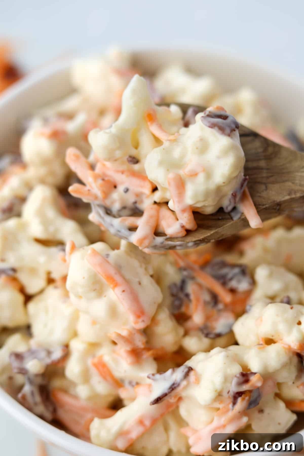 A wooden spoon lifting a generous portion of creamy cauliflower salad from a bowl, highlighting the texture and ingredients.