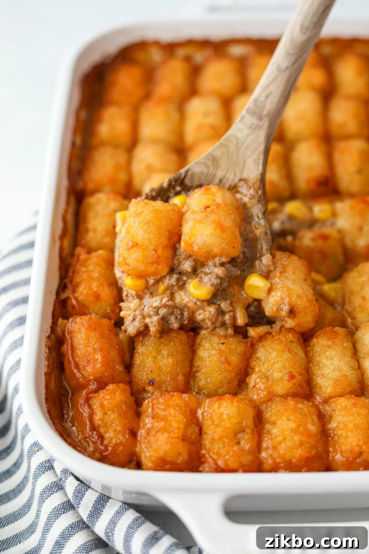A wooden spoon lifting a Tater Tot topping with some ground beef from a baking dish, showing the delicious layers.