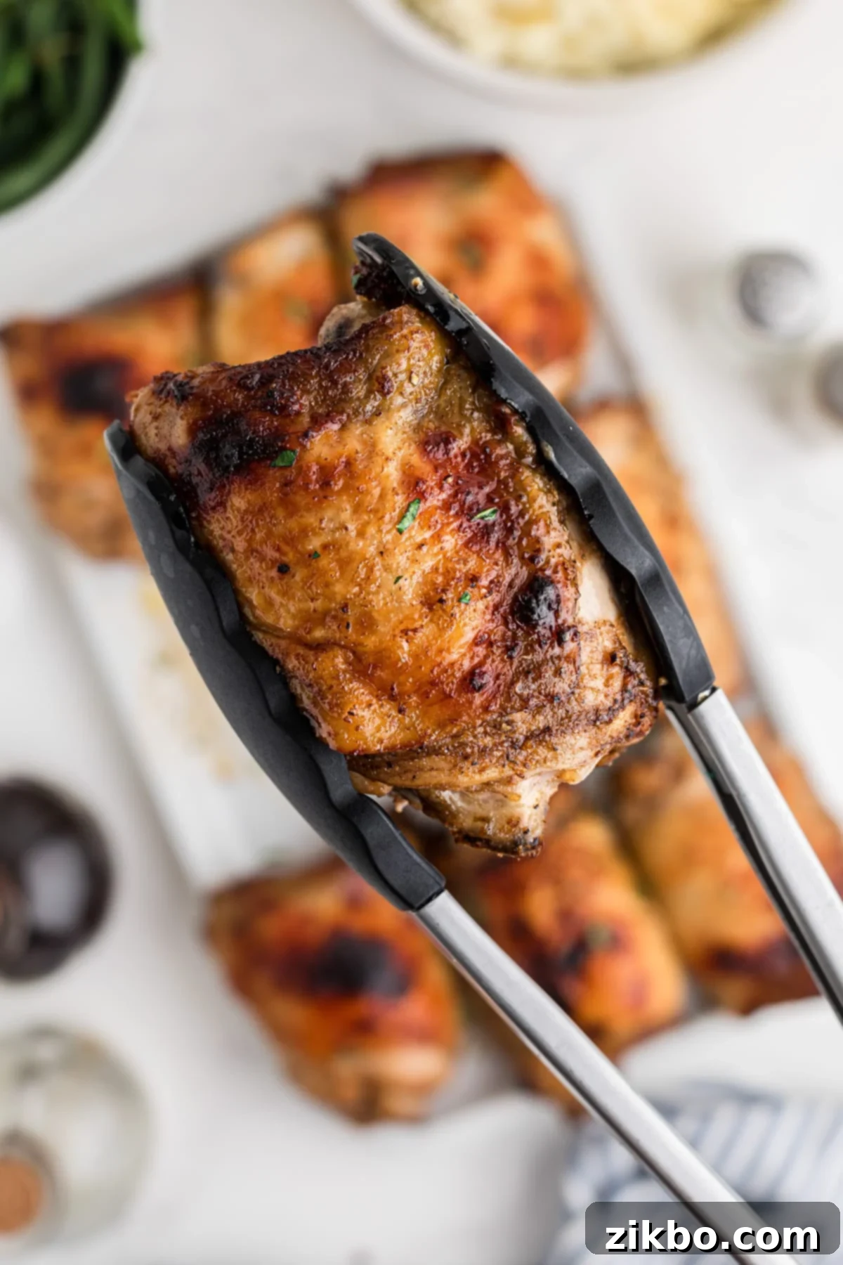 A pair of tongs lifting up a baked chicken thigh.