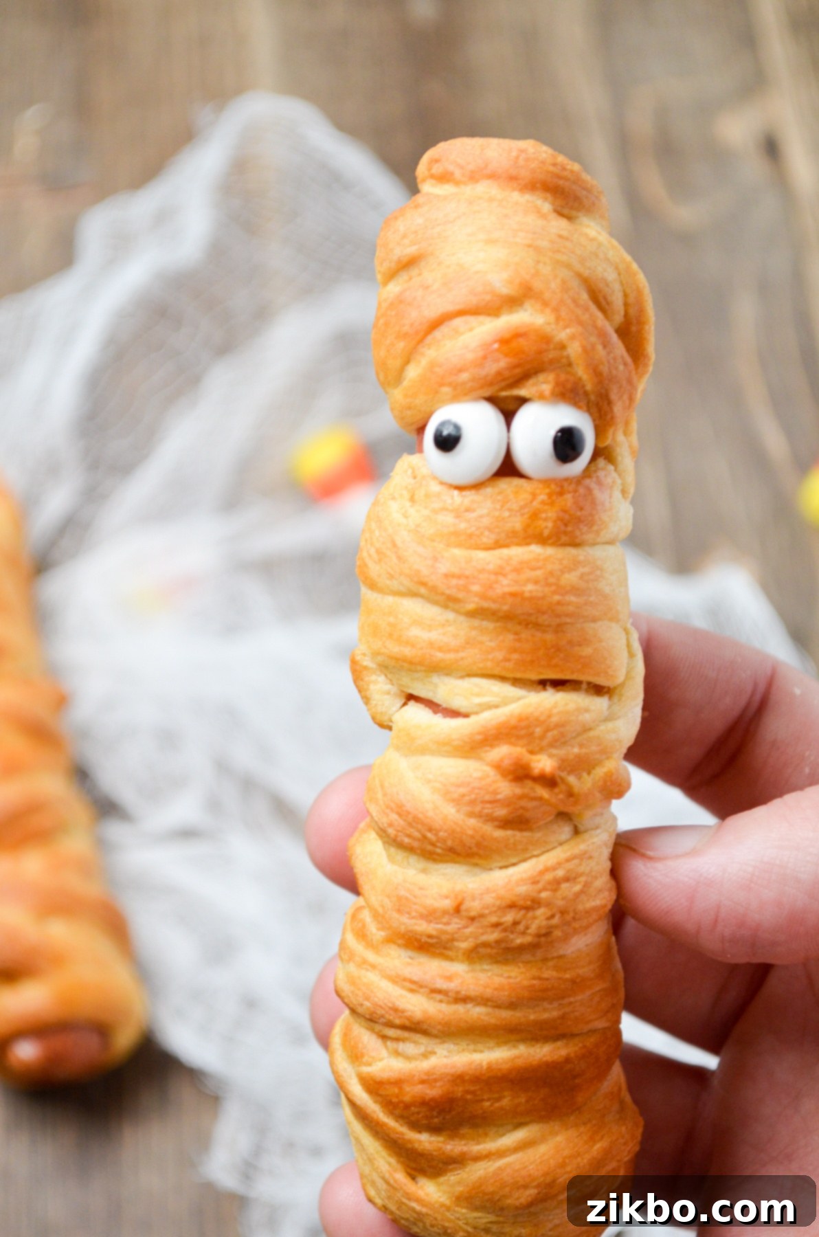 A mummy hot dog, with candy eyes fixed onto it, on a wooden board.