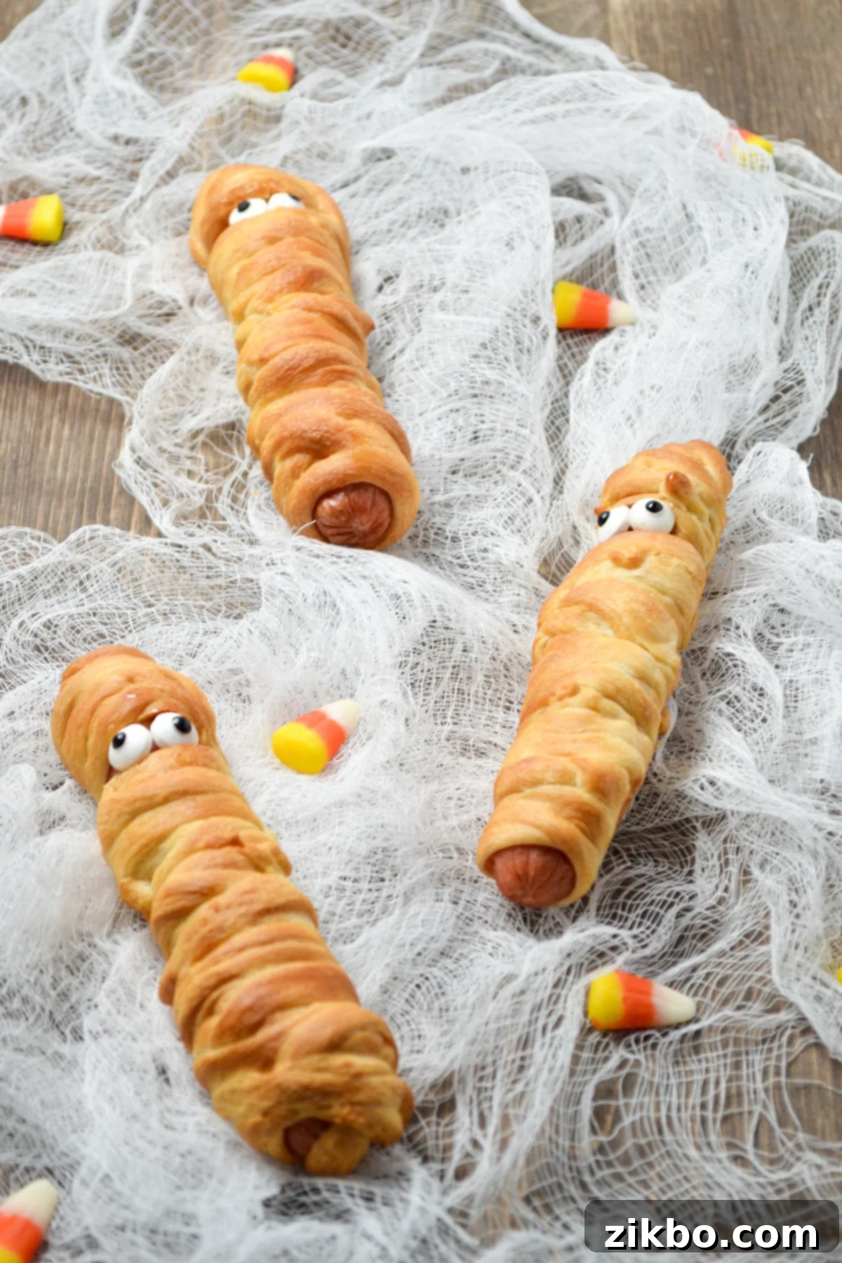 3 mummy hot dogs, laying on cheesecloth, garnished for Halloween.