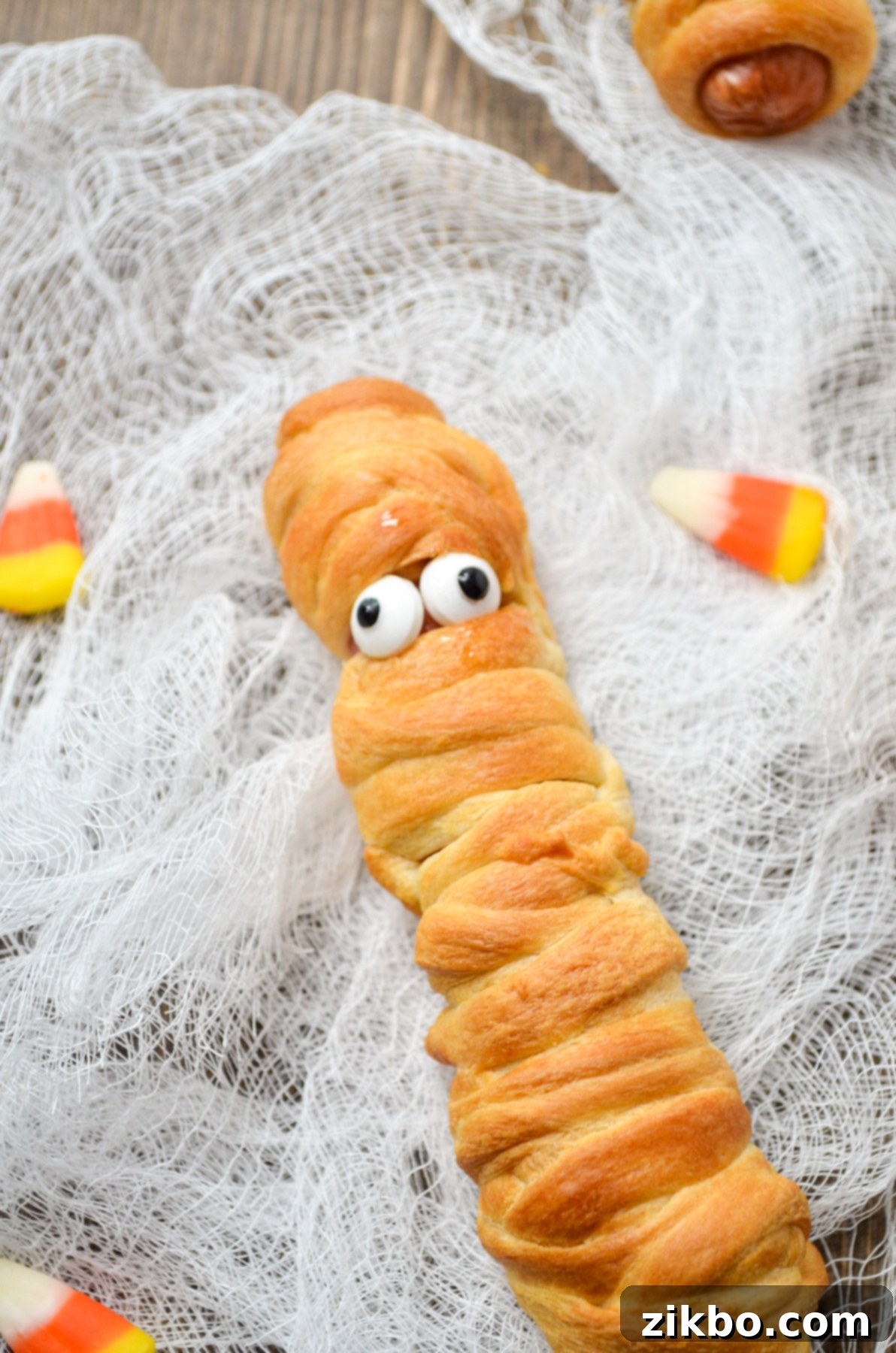 A hot dog wrapped in crescent roll dough, fitted with candy eyes, served with dipping sauce.