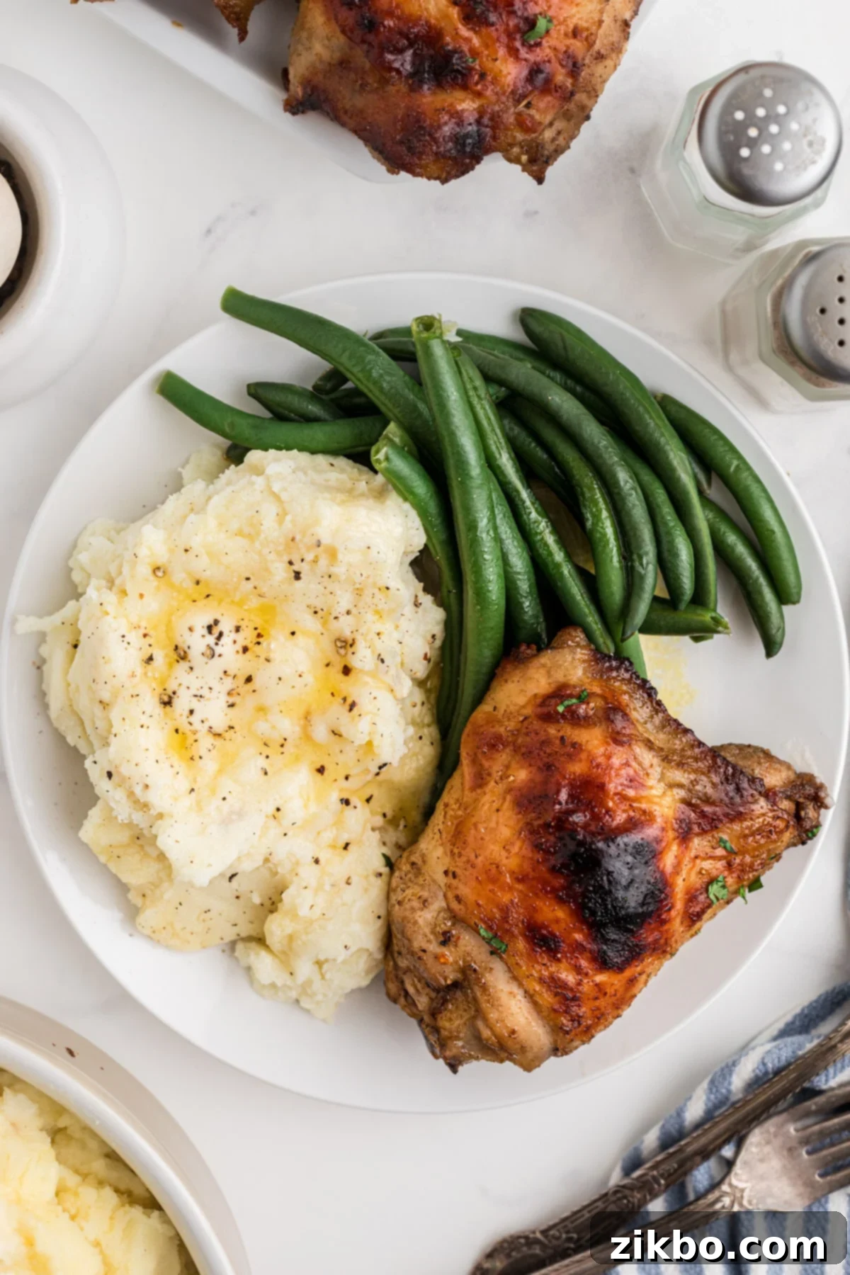 A plate featuring creamy mashed potatoes, fresh steamed green beans, and a perfectly crispy baked chicken thigh, artfully arranged.