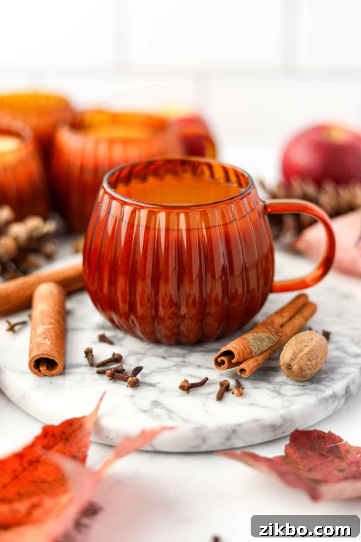 Golden Pumpkin Brew 7 An orange glass mug filled with pumpkin cider, with cinnamon sticks, cloves, and nutmeg artfully arranged around the glass on a marble surface.