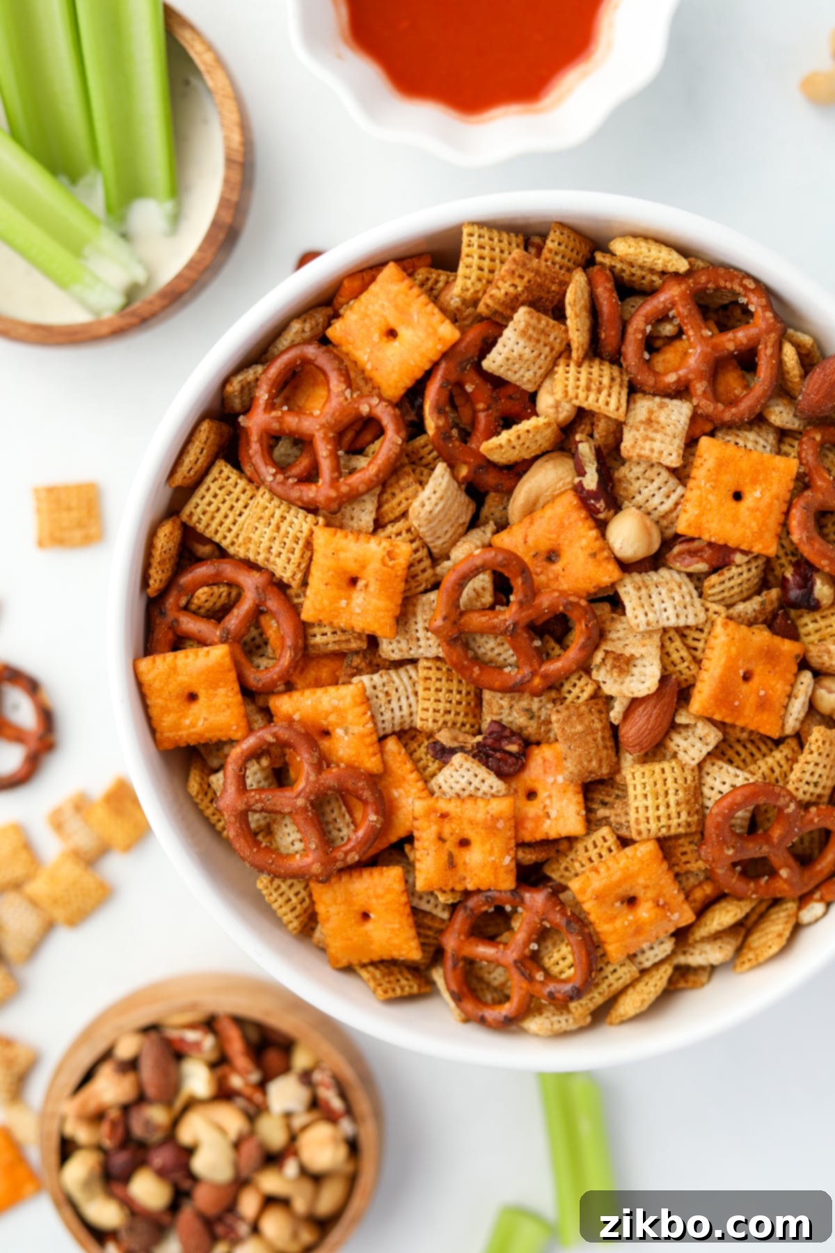 An overhead view of a bowl of Chex mix, with celery, nuts, and buffalo sauce in the background, ready to be enjoyed.