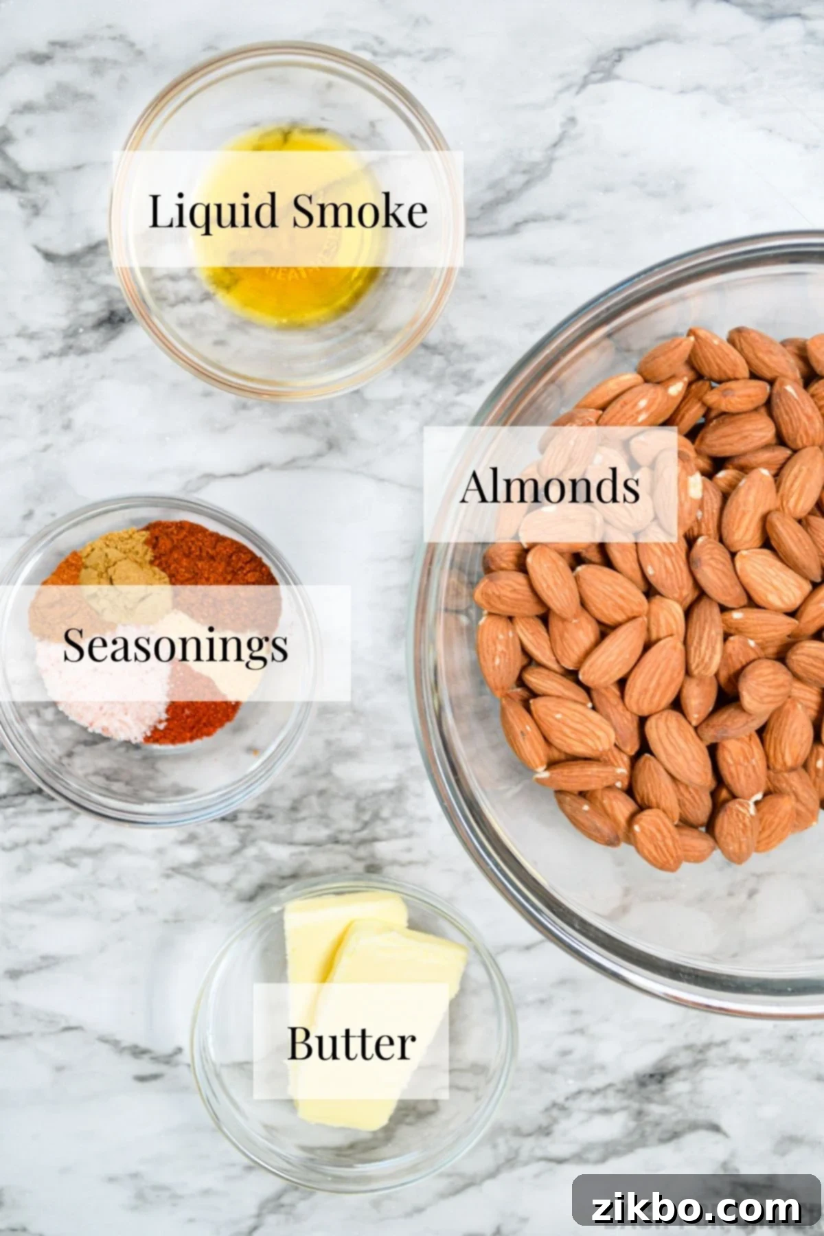 The ingredients needed to make roasted almonds: raw almonds, melted butter, liquid smoke, chili powder, cumin, garlic powder, cayenne, smoked paprika, and salt.